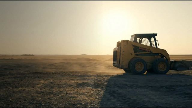 A wheeled skid steer in silhouette with the sun low behind it and dust in the air